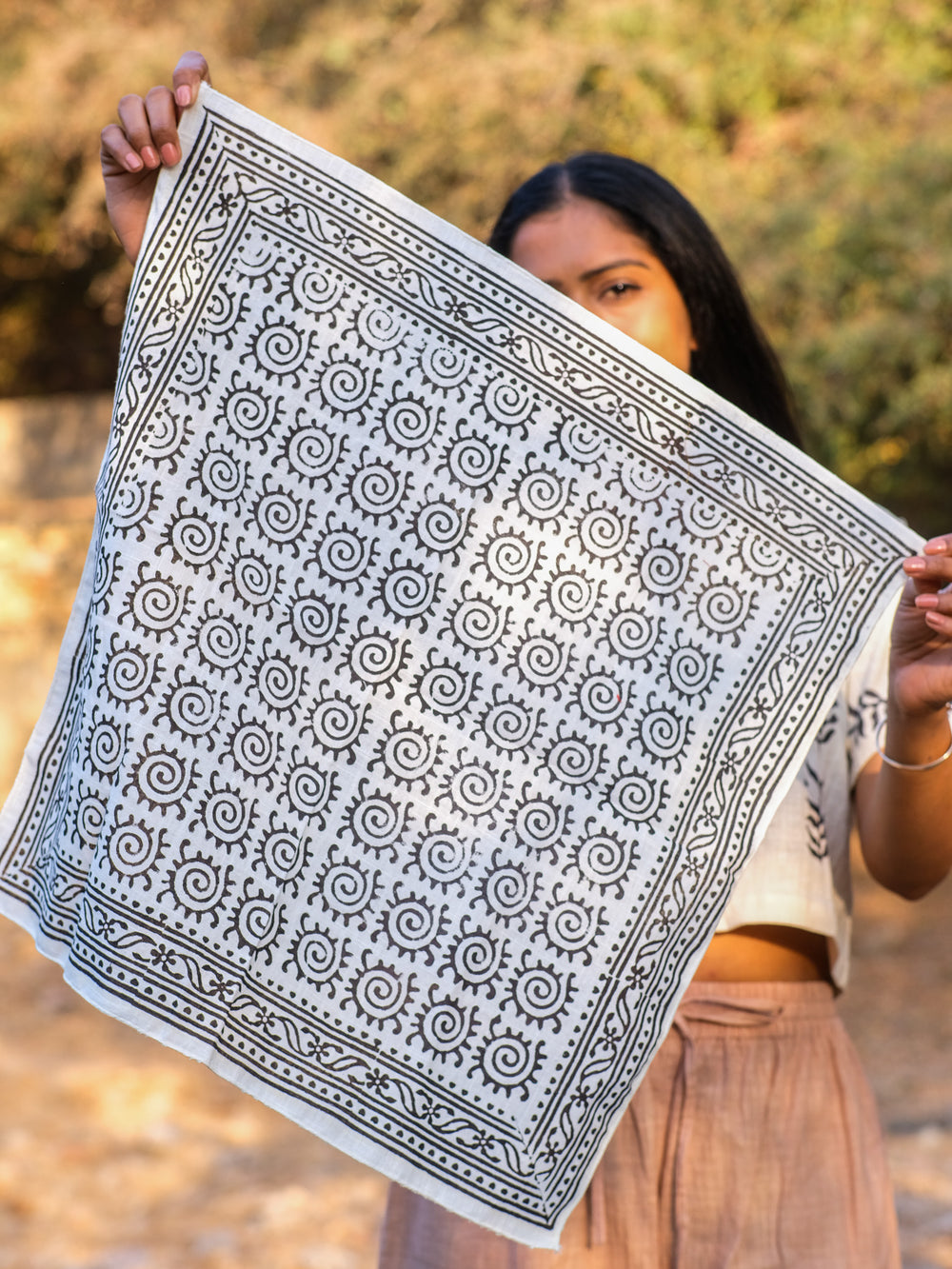 A person holds up the Presh Bandana, a square white accessory featuring intricate black geometric and spiral patterns, partially covering their face. Blurred trees and sunlight can be seen in the background.