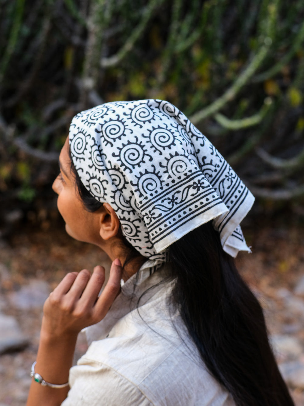 Wearing the Presh Bandana—a bohemian accessory with black patterns—a person with long dark hair and a white shirt sits outdoors, resting one hand near their chin, against a backdrop of blurred greenery.