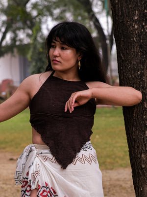 A woman with long dark hair stands near a tree, wearing a boho gypsy outfit. Her halter neck crop top, the Shanti Crop Top in dark brown, pairs with a patterned white wrap skirt. She gazes thoughtfully to the side with one arm resting on the tree trunk amidst lush greenery.