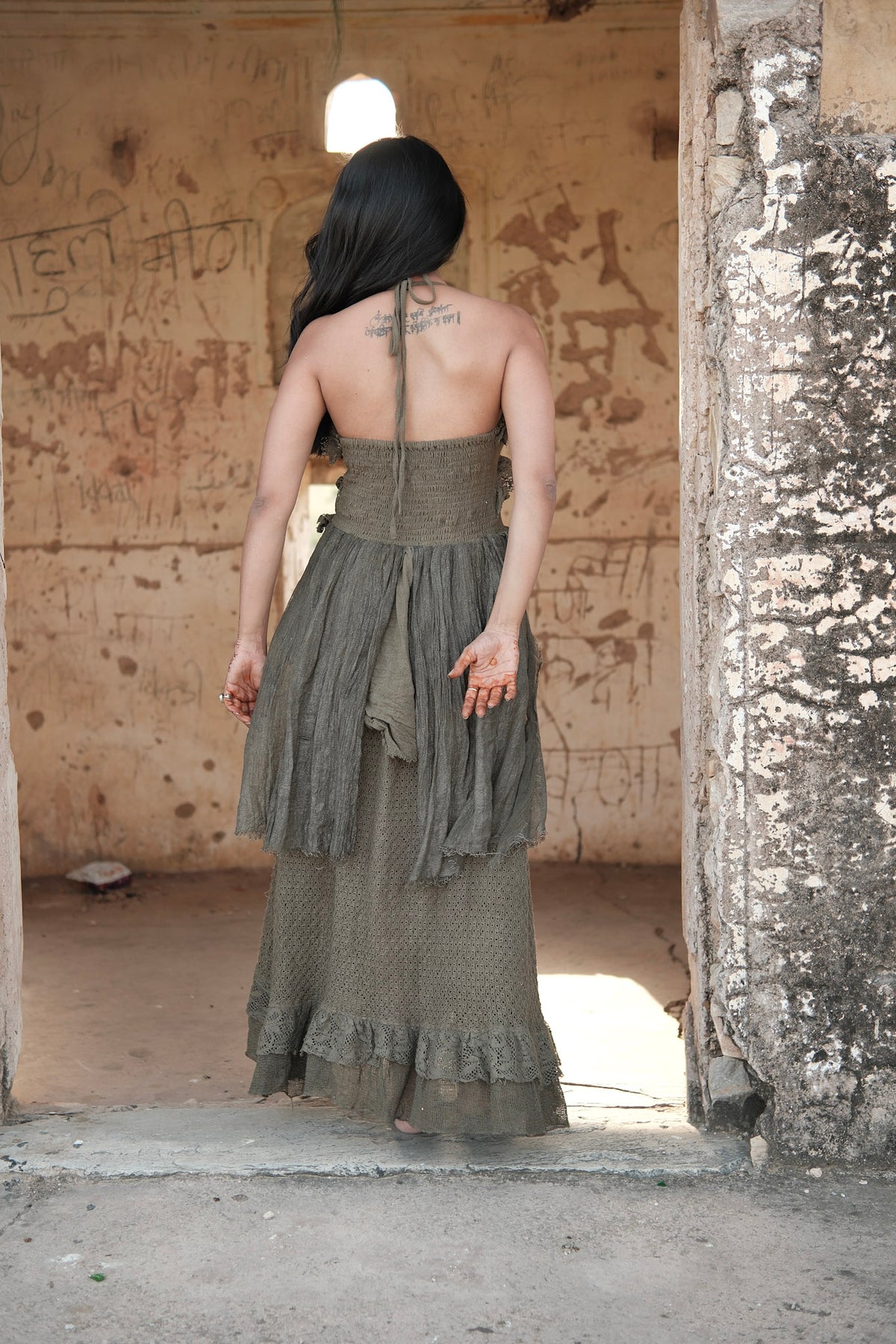 A person seen from behind wears the olive green Rust Ember Nomad Maxi Dress, a floor-length, bohemian halter-neck style with a textured bodice and ruffled hem, standing in the doorway of a weathered stone building.