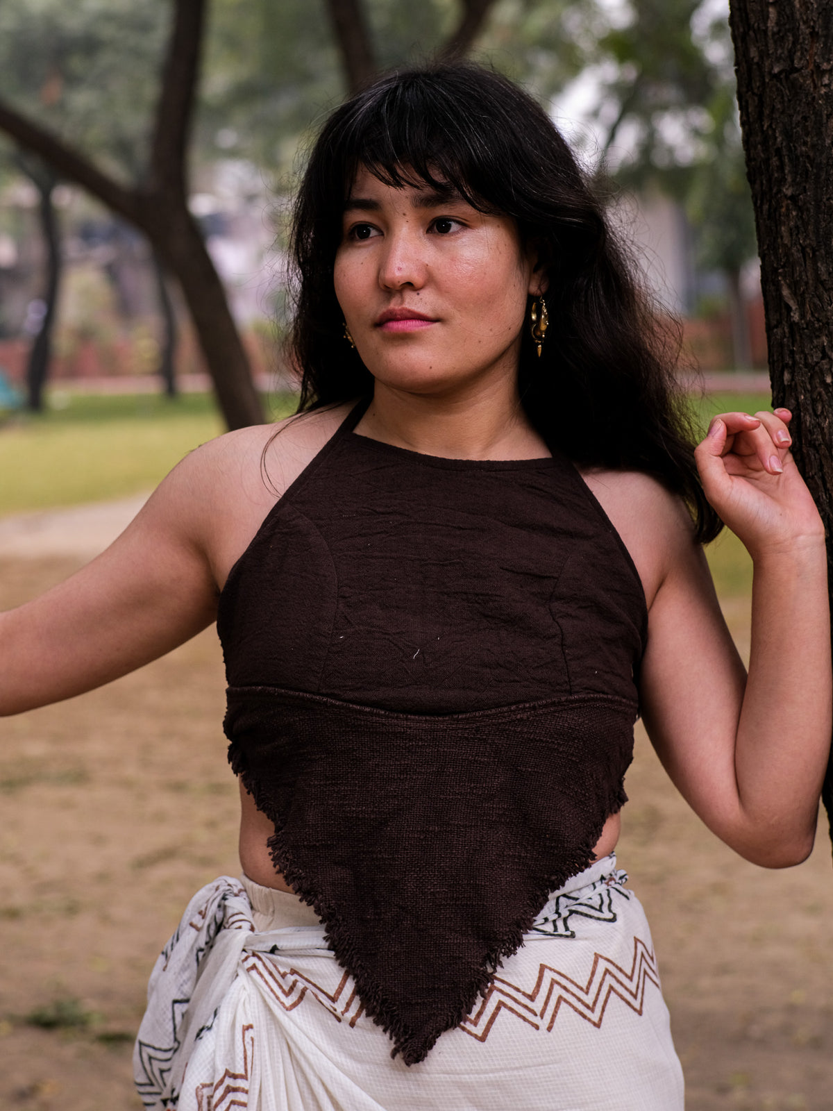 A woman with long dark hair stands outdoors wearing a Shanti Crop Top. Leaning against a tree with a contemplative look, she is enveloped by natures greenery, capturing the essence of boho gypsy style in her brown top and patterned wrap skirt.