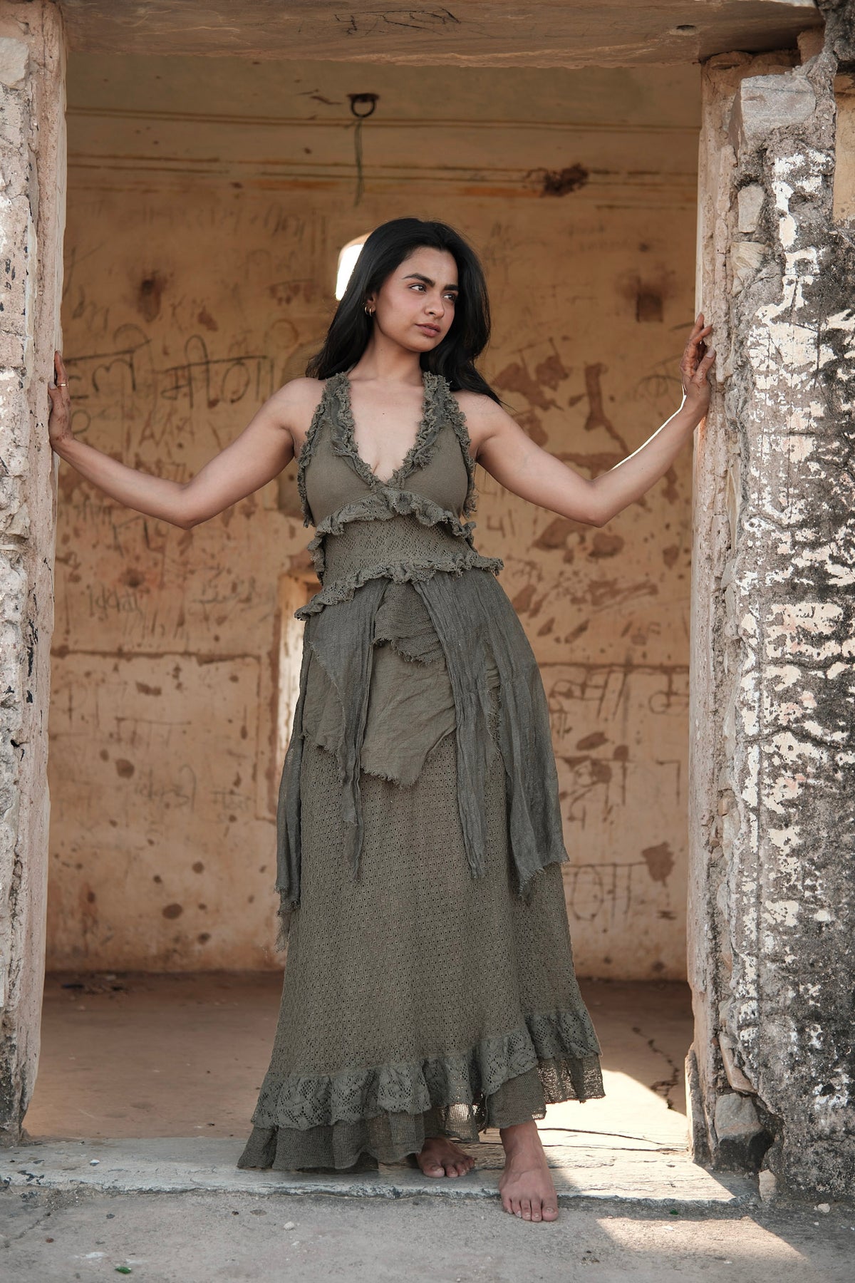 A woman wearing an olive green Rust Ember Nomad Maxi Dress with sleeveless, layered ruffled detailing standing in a stone doorway.