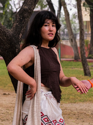 A woman stands outside near a tree, gazing at the camera in a brown Shanti Crop top with halter neck style, a boho gypsy skirt, and draped cloth on one shoulder. Grass and trees feature in the background as her left hand rests on her hip.