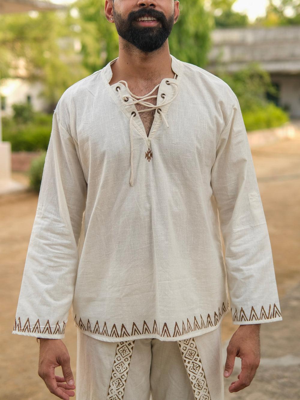 A man with a beard stands outdoors on a sunny day, wearing the Arok Kurta—a long-sleeved, cream artisanal kurta with matching pants, featuring intricate brown decorative patterns.