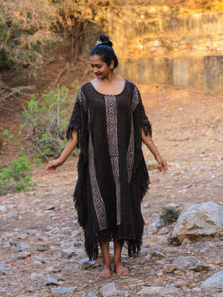 Barefoot on rocky terrain, a person drapes themselves in a Vama poncho with boho vertical stripes. With dark hair tied in a bun, they smile gently while looking down, set against dry foliage and a stone wall.