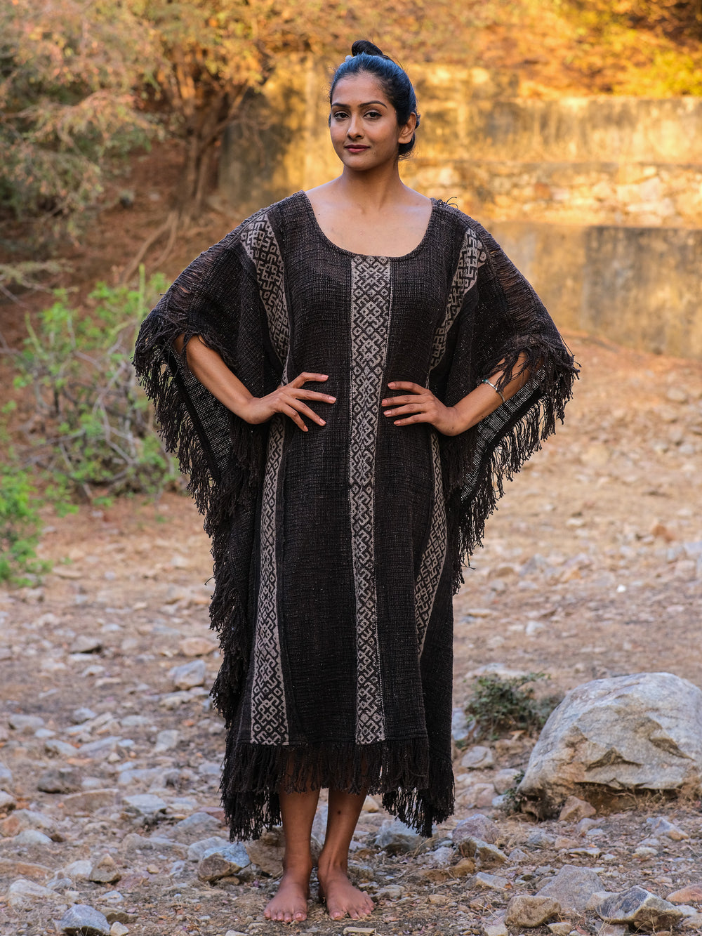 Standing confidently on rocky terrain, a person showcases their boho style with the Vama poncho, crafted from premium knitted fabric. The dark, fringed design with vertical stripes complements their short hair tied back as they rest their hands on hips amidst the dry ground and sparse vegetation.