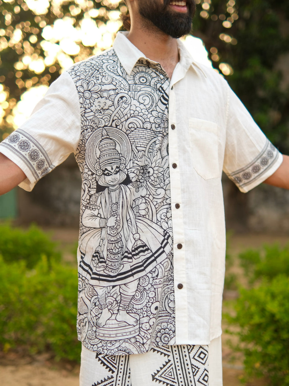 A man stands outdoors with arms open, wearing the Koothu Kathakali Shirt in a natural shade, crafted from breathable cotton and adorned on one side with black traditional Indian artistry featuring a Kathakali dancer.