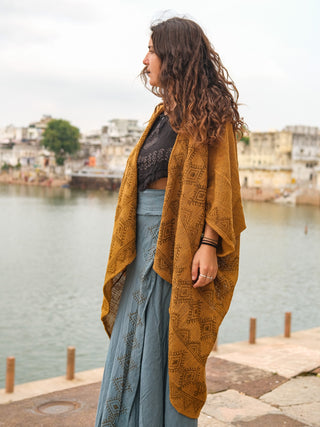 A woman with long, curly hair stands by the waterfront in a mustard yellow hand block-printed cotton Cocoon Shrug over a black top and blue skirt, with buildings and trees visible across the water.