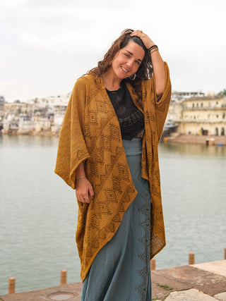 A smiling woman stands by water wrapped in a hand block-printed cotton Cocoon Shrug over a black top and blue-gray skirt, with buildings visible in the background.