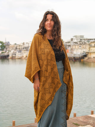 A woman with long wavy hair stands by a riverside, wearing a mustard yellow Cocoon Shrug over a black top and blue skirt. Buildings are visible on the opposite bank beneath a cloudy sky.