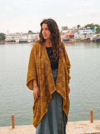 A woman with long curly hair, draped in the Cocoon Shrug—a hand block-printed mustard yellow shawl—over a blue skirt, stands by a calm waterfront with buildings and an overcast sky in the background.