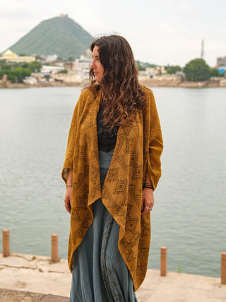 A woman with long curly hair stands by a waterfront, wearing a mustard-yellow hand block-printed cotton Cocoon Shrug and blue skirt, with a green hill and buildings in the background.