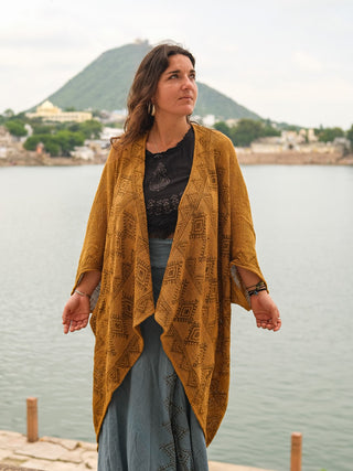A woman in a mustard yellow hand block-printed Cocoon Shrug and a long blue cotton skirt stands by a lakeside, hills and buildings in the background, gazing calmly to the side.