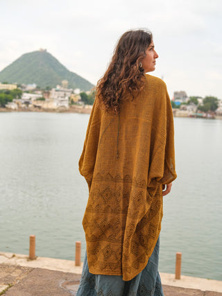 A woman with long curly hair, wearing the mustard yellow Cocoon Shrug and a blue cotton skirt, stands by a lakeside facing away, with hills and buildings in the background.