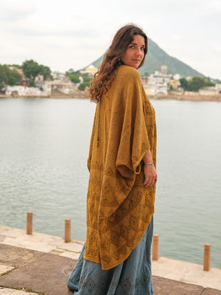 A woman with long wavy hair stands by a lake, looking over her shoulder, wearing a hand block-printed mustard cotton Cocoon Shrug and a blue skirt, with a town and mountain in the background.