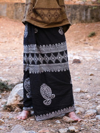 Close-up of a person wearing a patterned brown sweater and black-and-white Vasuki dhoti, standing barefoot on rocky ground, showcasing intricate geometric and paisley designs that evoke artistry against a blurred natural background, enhancing the scenes earthy charm.