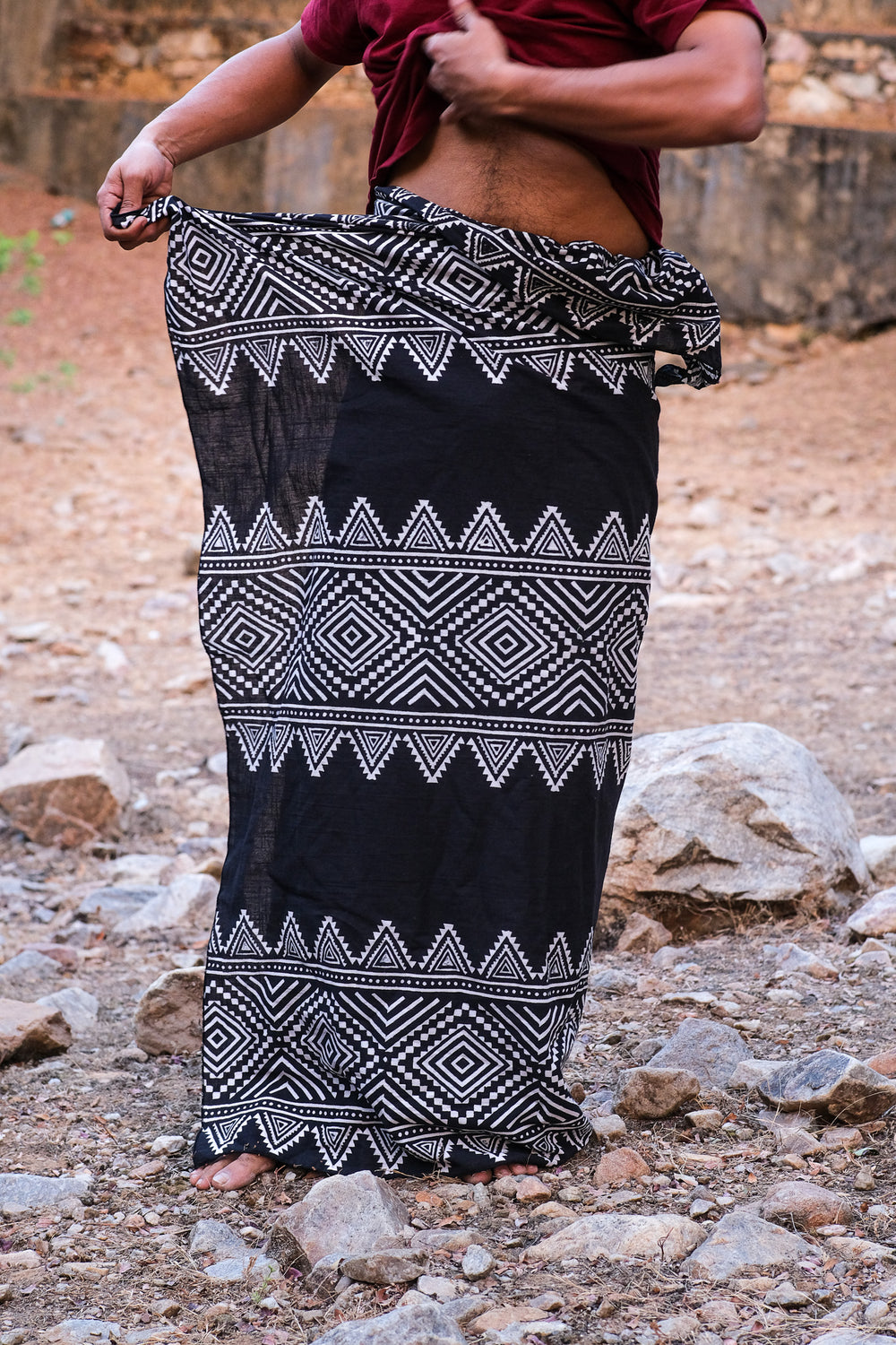 A person adjusts a black Vatsa Dhoti featuring a striking block print while standing outdoors on rocky ground, with their face not visible.