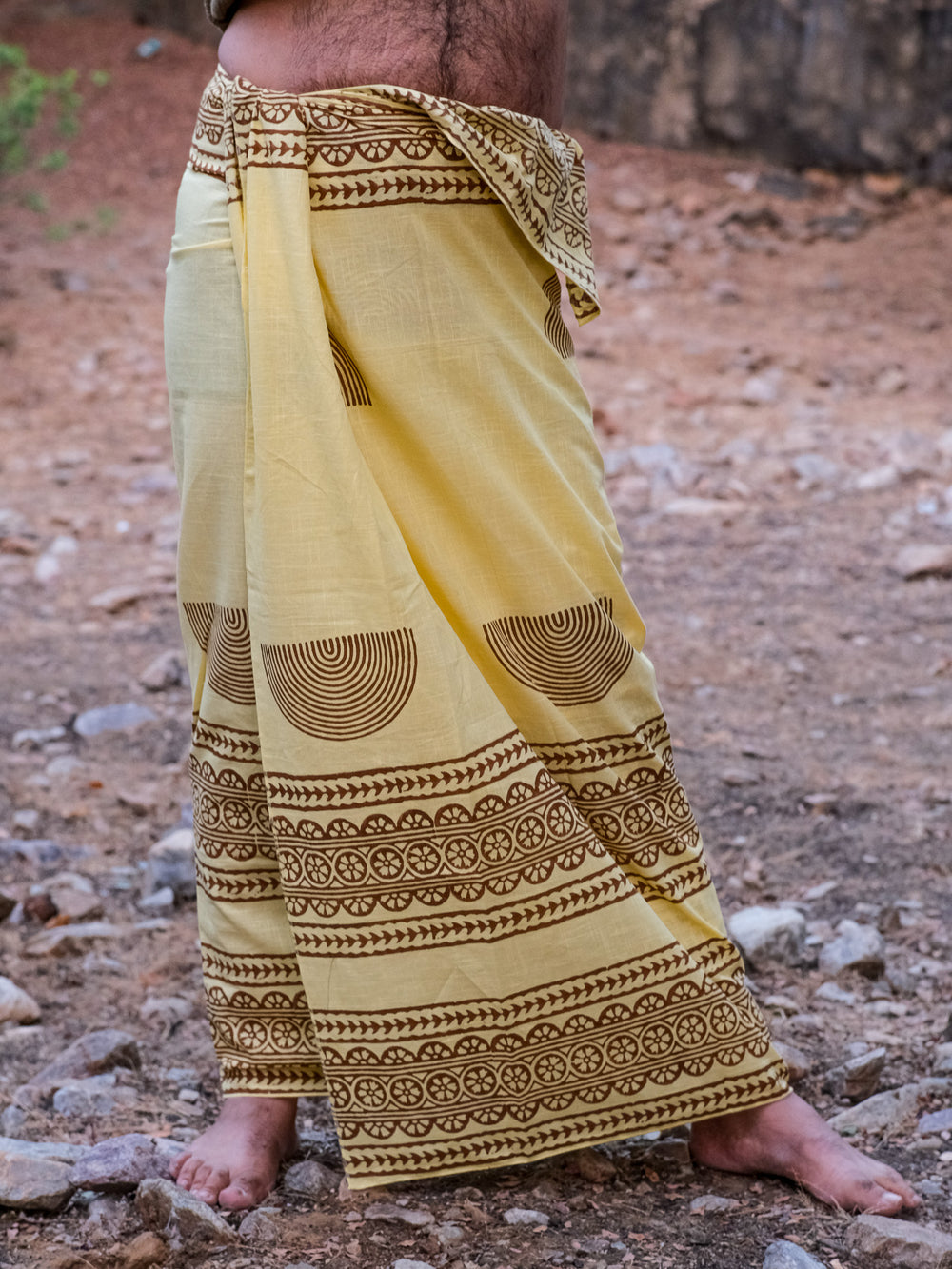 A person standing outdoors on rocky terrain, wearing the Urvi Dhoti in vibrant yellow with intricate brown patterns—a traditional Indian garment. Only their lower body and part of their torso are visible.