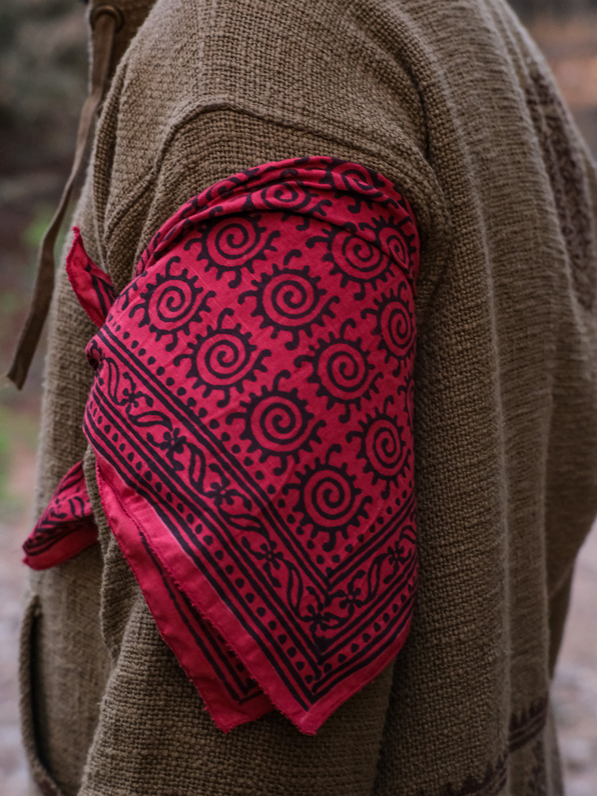 A person wearing a brown textured jacket has the red Presh Bandana tied around their upper arm, showcasing this versatile accessory in true bohemian style.