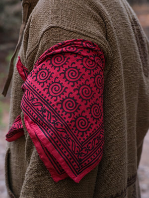 A person wearing a brown textured jacket has the red Presh Bandana tied around their upper arm, showcasing this versatile accessory in true bohemian style.