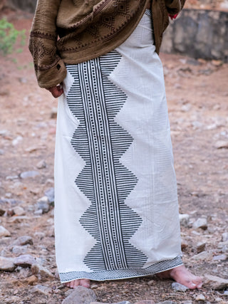 Outdoors, a person stands barefoot on rocky dirt, wearing the Anviksha Dhoti with black geometric block prints, elegantly paired with a brown long-sleeved top.