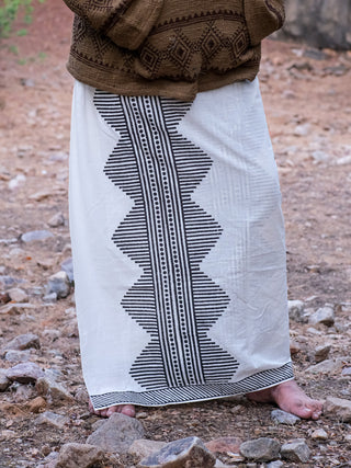 A person stands outdoors on rocky terrain, wearing an Anviksha Dhoti with black geometric block prints. They are barefoot and paired with a snug brown patterned sweater.