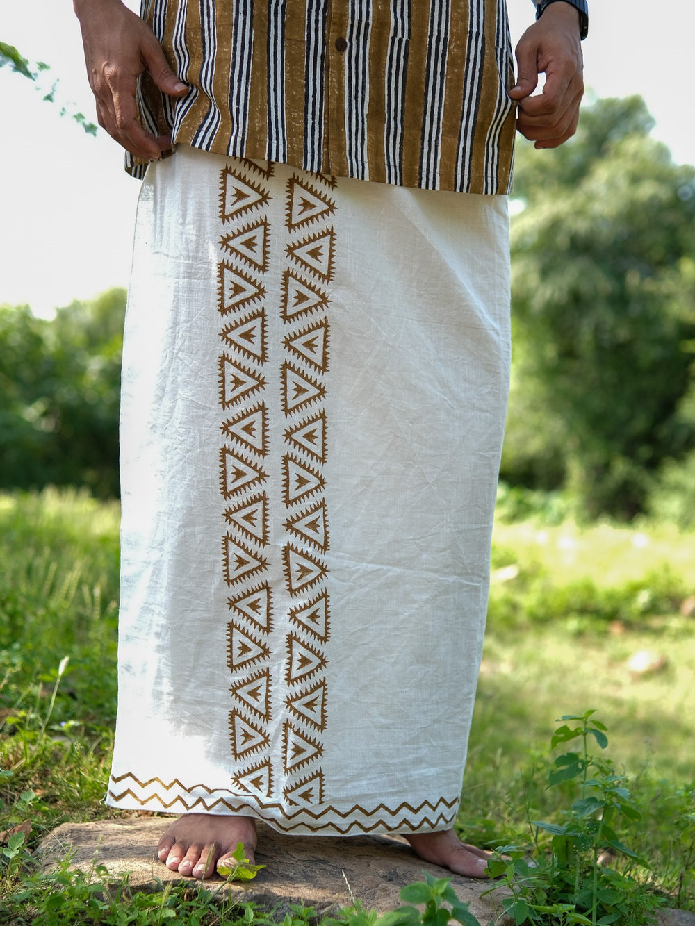 A person stands barefoot on grass, wearing the “morya dhoti”—a white cotton garment featuring brown geometric Rajasthani handblock patterns—and a striped shirt. The background is green and outdoors.