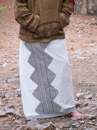 A person stands barefoot on rocky ground, wearing a brown sweater with geometric patterns and the Anviksha Dhoti, a long white skirt featuring black zigzag stripes reminiscent of traditional block print.