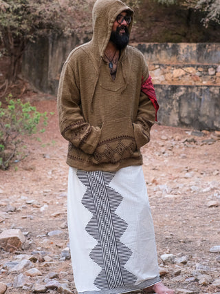 A person in a brown hooded sweater with a red armband and Anviksha Dhoti, a long white skirt with black geometric patterns inspired by traditional Indian attire, stands outdoors in a rocky setting. They are wearing glasses.