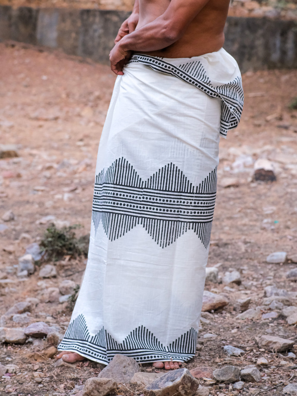Wearing the Arya Dhoti in white with a black block print, a barefoot person stands on rocky ground, showing only their lower torso and legs.