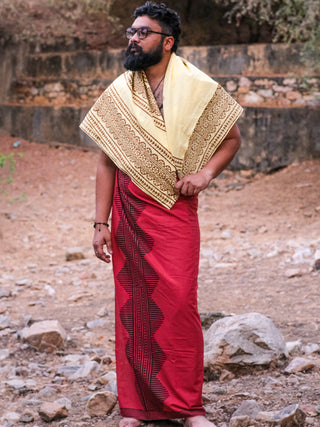 A person in glasses and traditional Indian attire, including the red Anviksha Dhoti and a yellow and black shawl, stands on rocky ground with a stone wall and trees behind them, exuding timeless elegance.