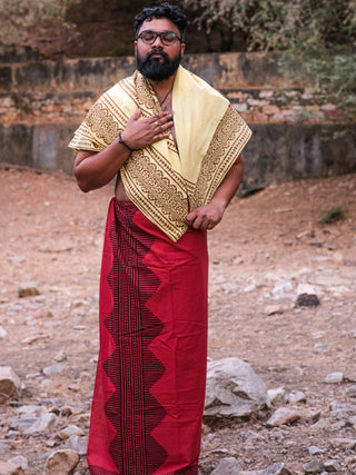 A man stands on rocky terrain, wearing glasses and showcasing traditional Indian attire. He drapes a patterned cream shawl over his shoulder and wraps a red Anviksha Dhoti with black block print designs around his waist while holding the shawl to his chest.