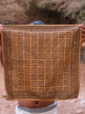 A person holds up the Presh Bandana in Camel Brown, featuring a repeating black spiral pattern and decorative border, obscuring their face and upper body while a natural outdoor background is visible.