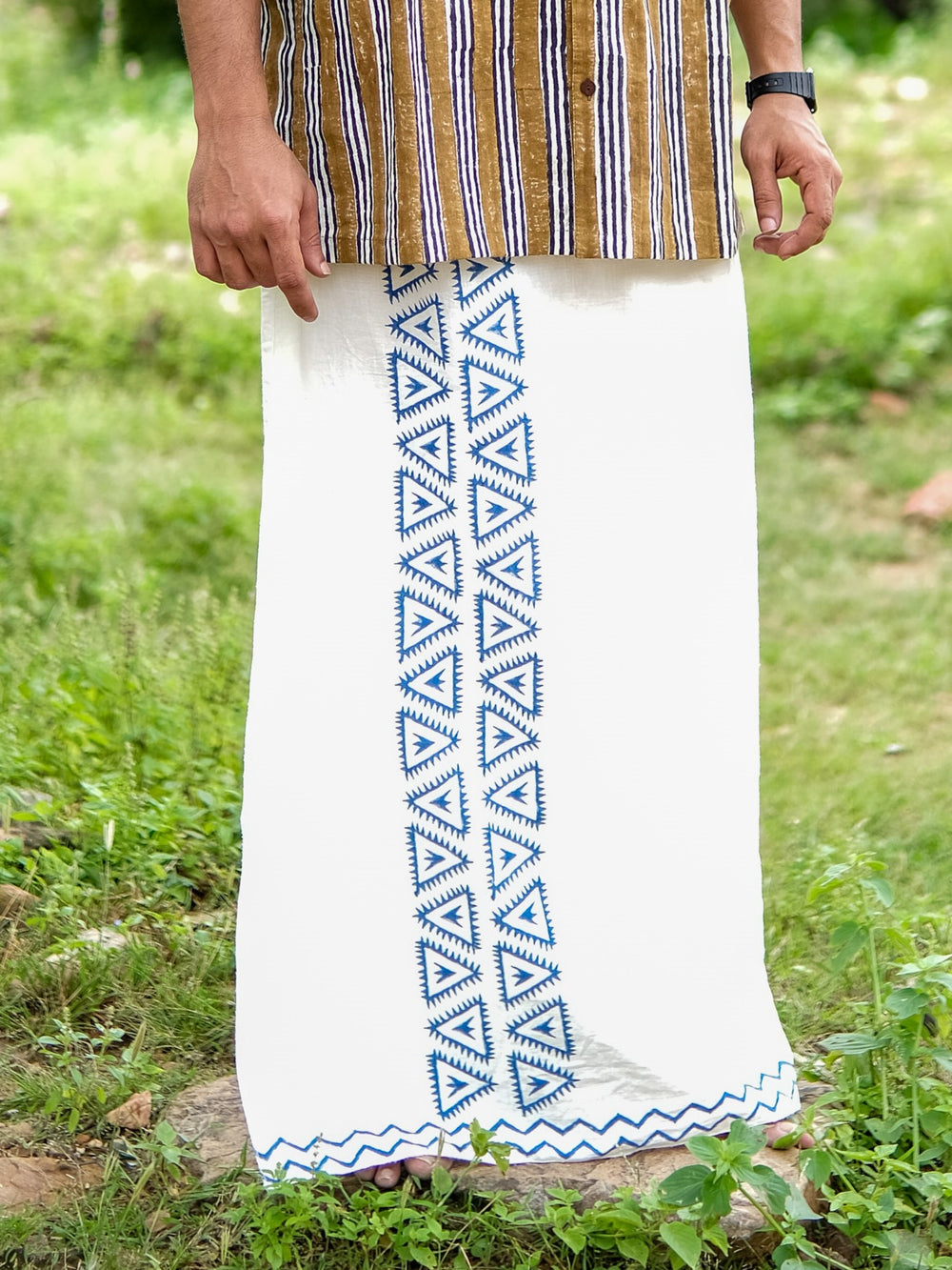 A person stands on grass wearing the morya dhoti, a white cotton garment featuring a vertical blue geometric pattern, paired with a striped shirt and a wristwatch. Only the lower body is visible.