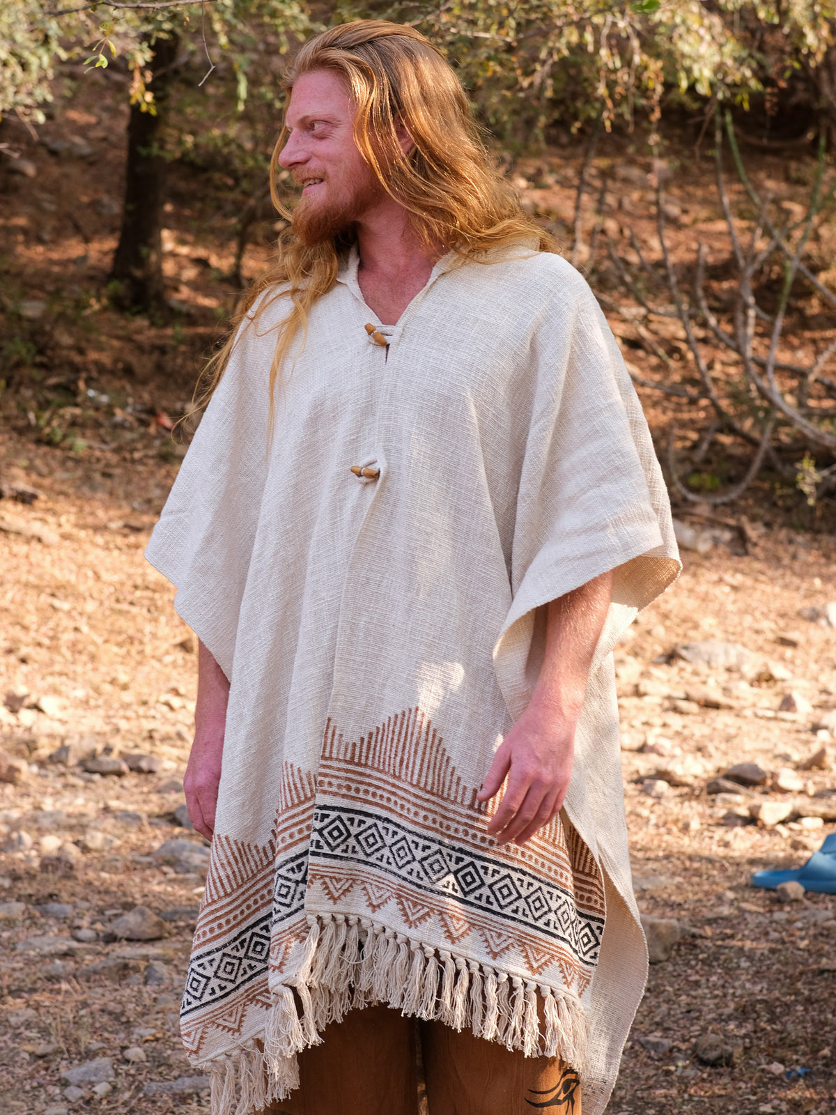 A person with long red hair stands outdoors, smiling and looking to the side while wearing a cream-colored poncho with geometric patterns and fringe, elegantly paired with the Swing Shawl draped over their shoulders.