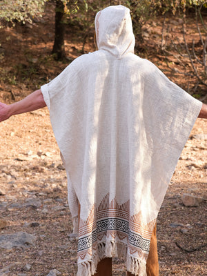 A person stands outdoors with arms outstretched, facing away from the camera, wearing a hooded cream-colored poncho with patterned trim, their look elegantly complemented by the Swing Shawl draped over their shoulders.