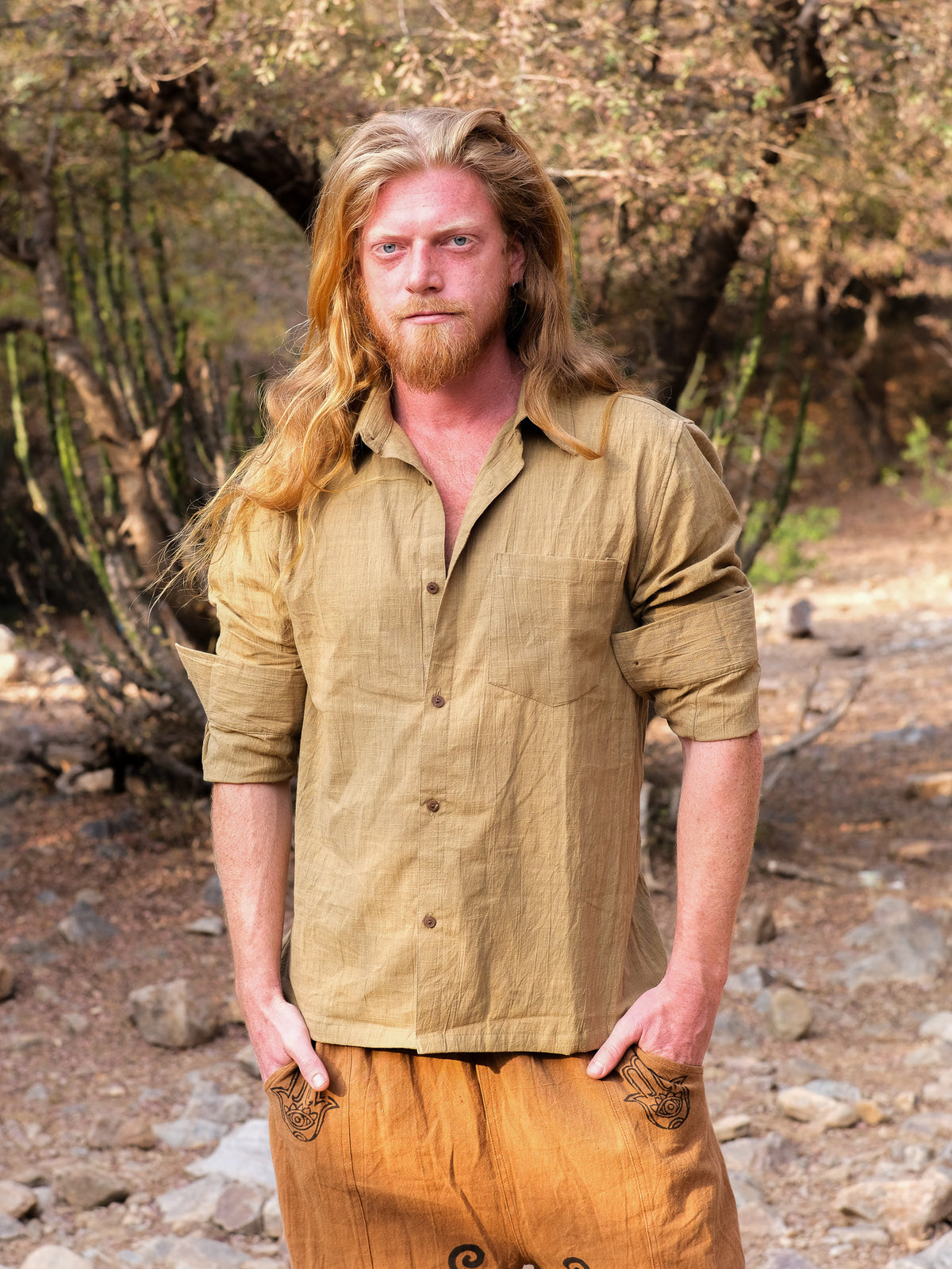 A man with long blond hair and a beard stands outdoors wearing the YinYang Shirt in sandyellow and light brown pants, with trees and rocks in the background.