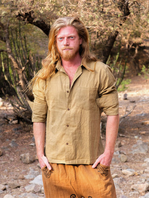 A man with long blond hair and a beard stands outdoors wearing the YinYang Shirt in sandyellow and light brown pants, with trees and rocks in the background.