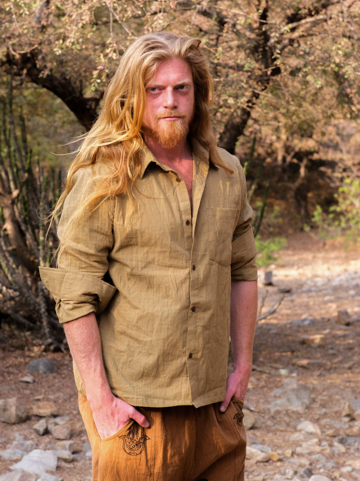 A man with long blond hair and a beard stands outdoors in a natural, rustic setting of trees and rocks. He is wearing sandyellow YinYang Shirt and matching pants, the shirt’s subtle design adding an element of balance to his earthy ensemble.