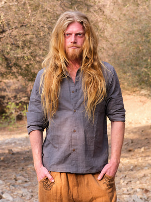 A person with long blond hair and a beard stands outdoors, wearing the gray YinYang Shirt and brown pants, with hands in pockets and a natural, blurred background.