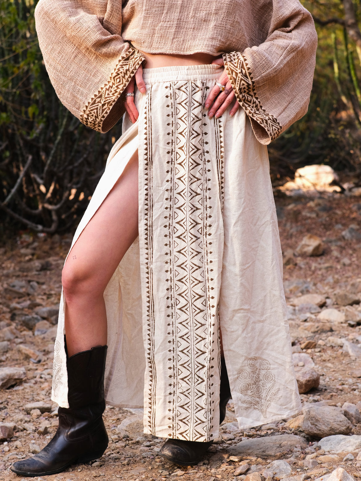 Full-length front view of the model in the cream beige cotton block print wrap skirt with a deep side slit, showing the hand printed geometric centre panel and the floral motif at the hem, worn with a cream blouse and black cowboy boots.
