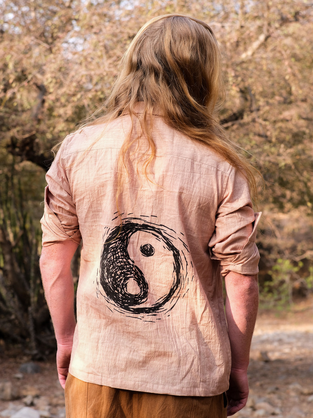 A person with long blond hair stands outdoors, facing away, wearing the beige YinYang Shirt that features a large, hand-drawn yin-yang symbol on the back. Trees and rocks can be seen in the background.