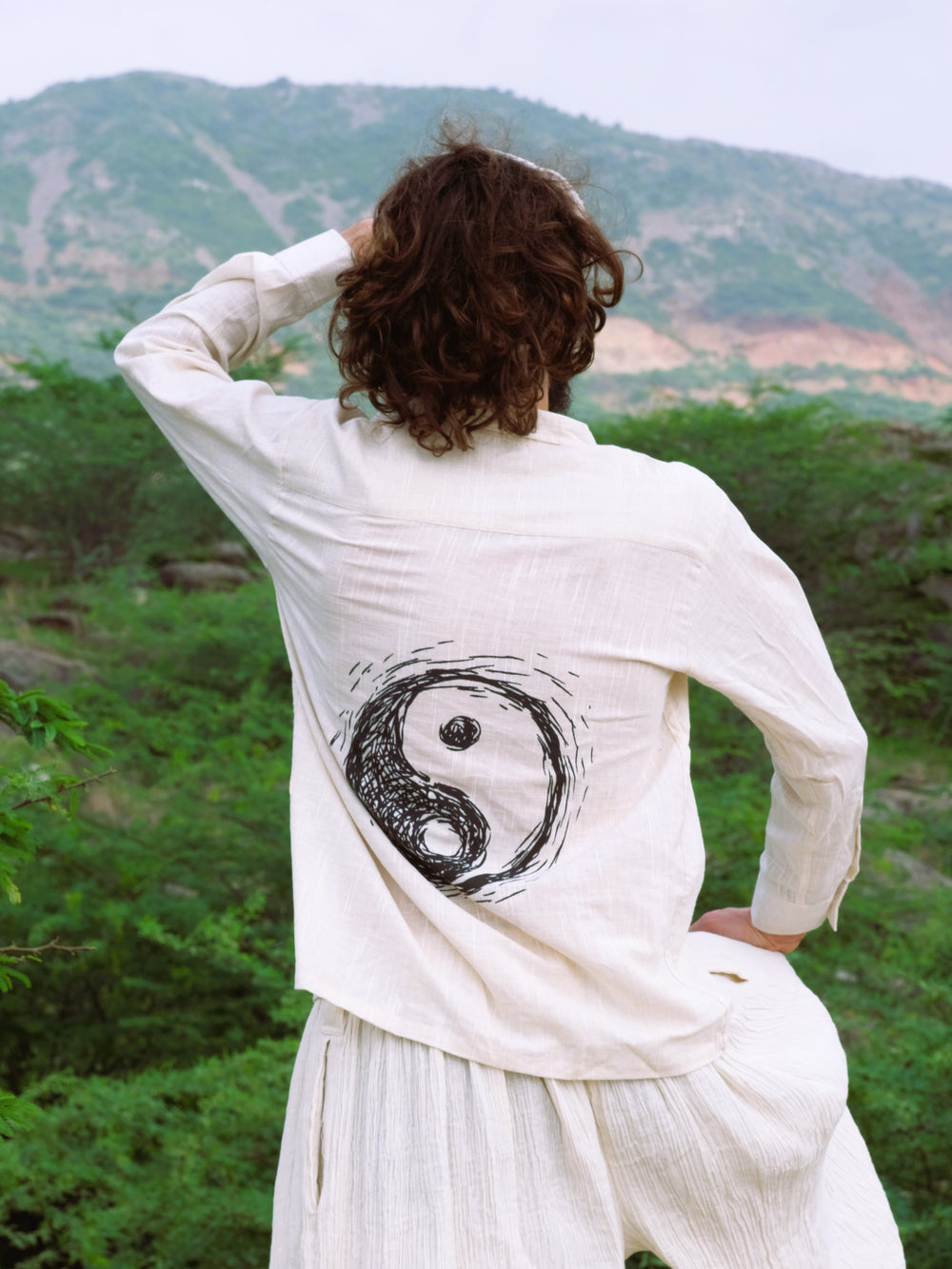 A person with curly hair, wearing a natural-colored YinYang Shirt with a large yin-yang symbol on the back, stands outdoors overlooking a green landscape and hills.
