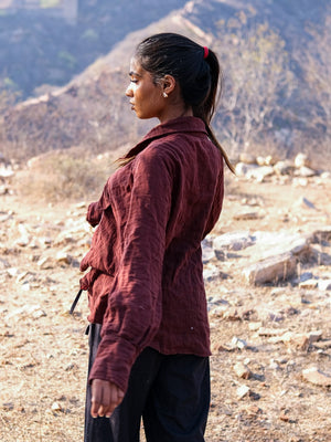 A woman with a ponytail, wearing a brown jacket and black pants, stands outdoors on rocky, dry terrain, gazing to her left. Her relaxed silhouette is complemented by the effortless style of the Ciraya Baggy Shirt layered beneath her jacket.