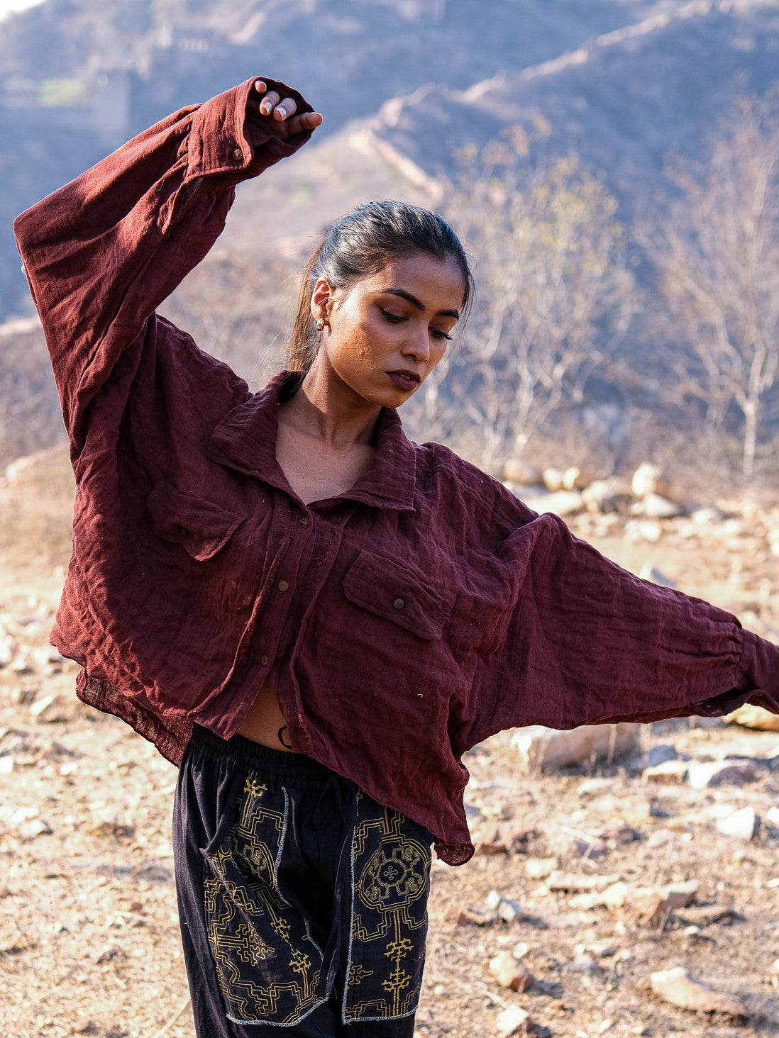 A woman stands outdoors on rocky terrain, raising one bent arm, wearing a loose maroon Ciraya Baggy Shirt paired with black pants. Dry trees and hills form the background.