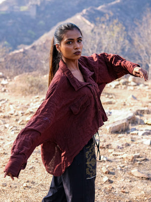 A woman with long hair poses outdoors against rocky terrain and hills, wearing a loose maroon Ciraya Baggy Shirt paired with black pants.