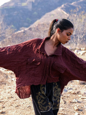 A woman stands outdoors with her arms outstretched in front of rocky terrain and distant hills, wearing the Ciraya Baggy Shirt in maroon and black patterned pants.