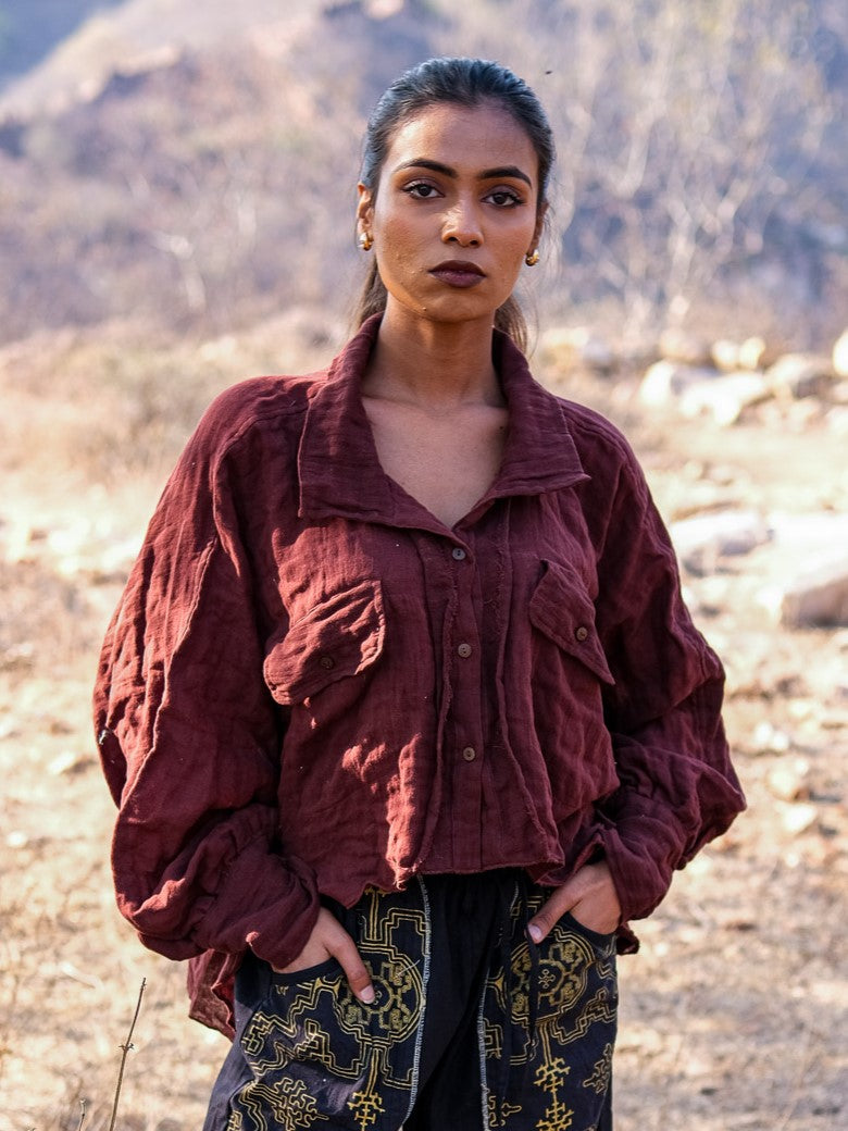 Outdoors amidst dry grass and rocks, a woman stands confidently with her hands in her pockets, wearing a maroon Ciraya Baggy Shirt featuring balloon sleeves and patterned black pants. The background is softly blurred as she faces the camera.