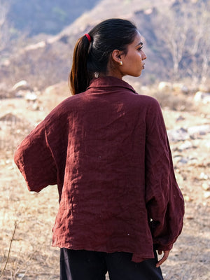 A woman with a ponytail, dressed in a loose maroon Ciraya Baggy Shirt, stands outdoors with her back to the camera and glances to the side, with dry grass and rocky hills in the background.