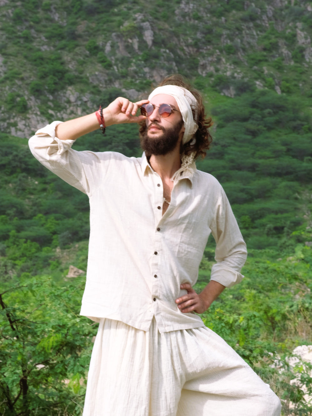 A man wearing the YinYang Shirt and sunglasses poses with one hand on his hip and the other adjusting his glasses, standing outdoors against a lush green hillside.
